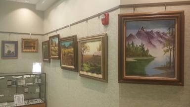 Photo of several paintings along wall. Foremost is a lake with mountains.