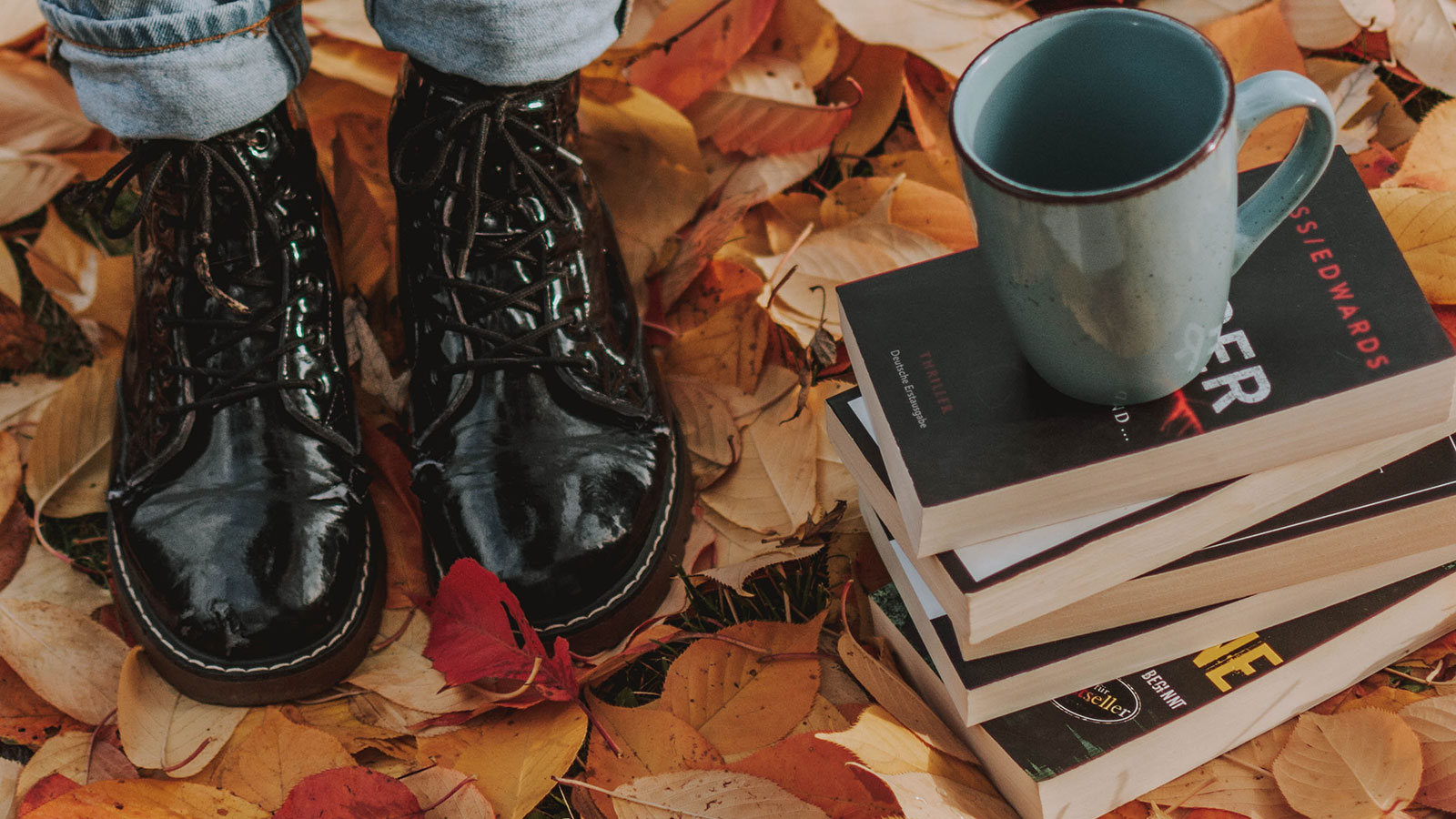 Closeup of person in shiny black boots standing next to books on fall leaves.